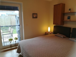Quarto com cama, mesa de cabeceira, luminária, janela com vista para um pátio e um pequeno quadro na parede.