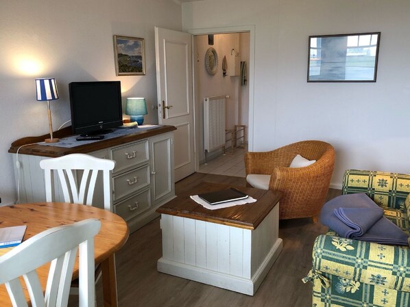 A living room with a wooden table, a white chair, a wicker armchair, and a patterned sofa. A television is on a cabinet, and there is a door leading to another room.