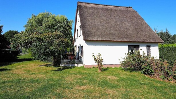 Une maison blanche avec un toit de chaume, entourée d’une pelouse verdoyante et d’arbres.