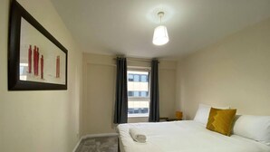 Ein Hotelzimmer mit Bett, Nachttisch, Lampe, einem Fenster mit Vorhängen und einem gerahmten Bild an der Wand.