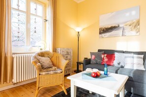 A cozy living room with a wicker chair, a grey sofa, a white coffee table with apples, and a lamp.