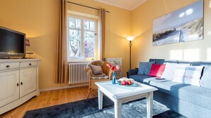 A living room with a television, a sofa, a chair, a table with fruit, a lamp, and a window with curtains. - Neue Reihe 22 Whg. 1 "Villa Balticum" - Modern vacation apartment close to the center (Zingst)
