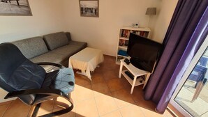 En stue med sofa, stol, lille bord, fjernsyn på et stativ og reol.