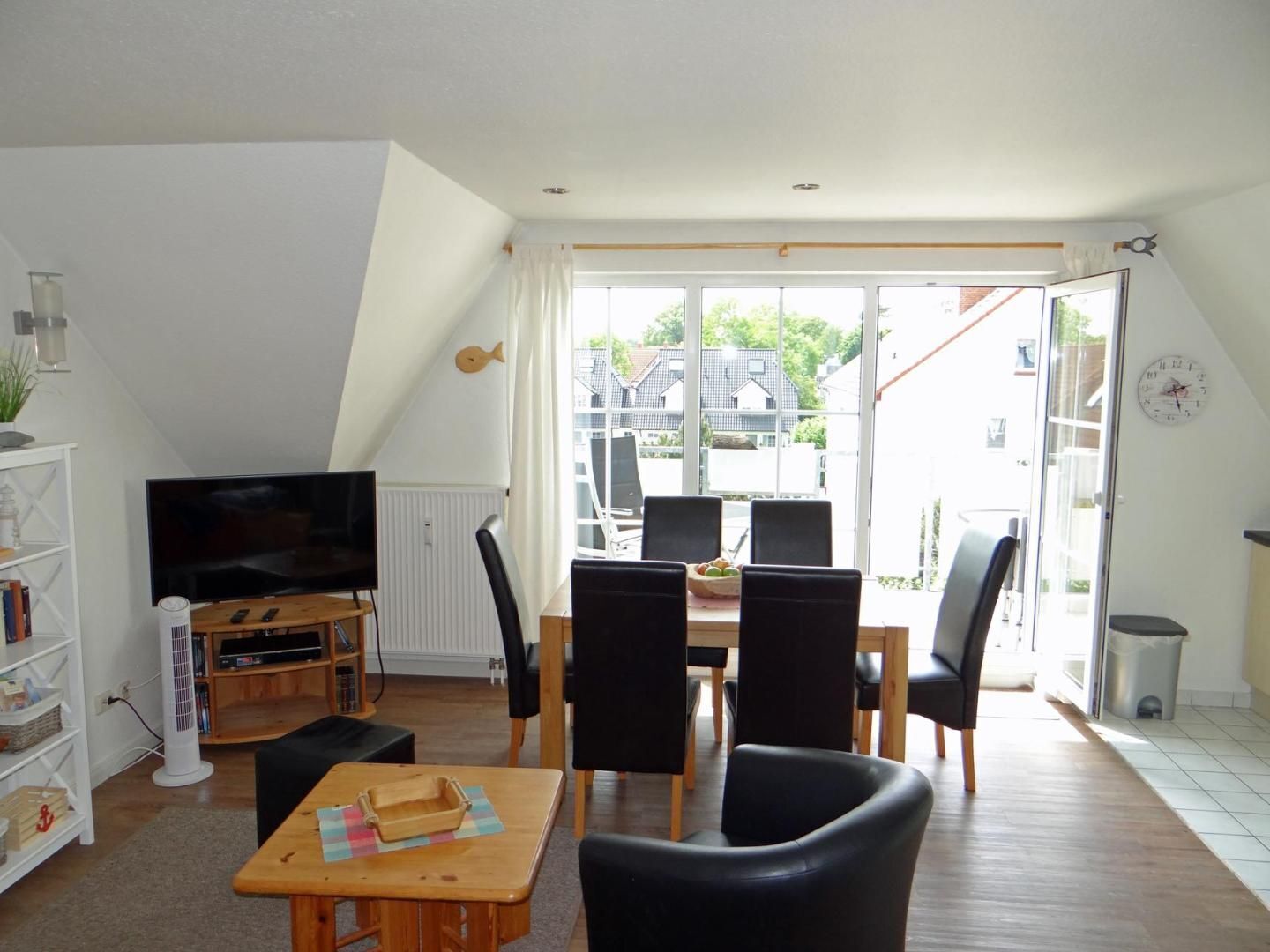 Uma sala de estar com televisão, uma mesa de jantar com cadeiras, uma estante de livros e uma janela grande com vista para casas.