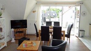Una sala de estar con televisor, una mesa de comedor con sillas, una estantería y un ventanal con vistas a casas.