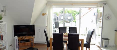 Uma sala de estar com televisão, uma mesa de jantar com cadeiras, uma estante de livros e uma janela grande com vista para casas.