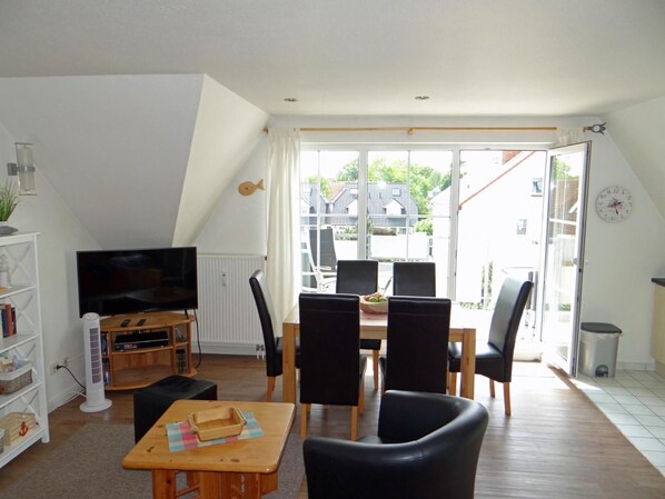 A living room with a TV, a dining table with chairs, a bookshelf, and a large window with a view of houses. - Strandstrasse 51, Apt. 10 — comfortable vacation apartment in a building (Zingst)