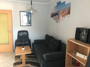 A living room with a black sofa, a wooden coffee table with a plant, a black leather chair, a bookshelf with books, and a mounted picture of a beach scene.