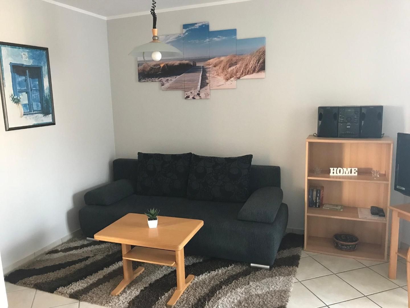 Ein Wohnzimmer mit einem dunkelgrünen Sofa, einem Holztisch, einer kleinen Pflanze, einem Gemälde mit Strandmotiv, einem Bücherregal mit der Aufschrift „HOME“ und einem Fernseher auf einem Standfuß.
