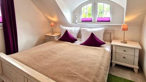 A bedroom with a sloped ceiling, a bed with a beige cover, purple pillows, two bedside tables with lamps, and a window with a view of greenery.