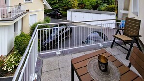 A balcony with a table and chairs, a car parked in the background, and a view of neighboring houses and trees.