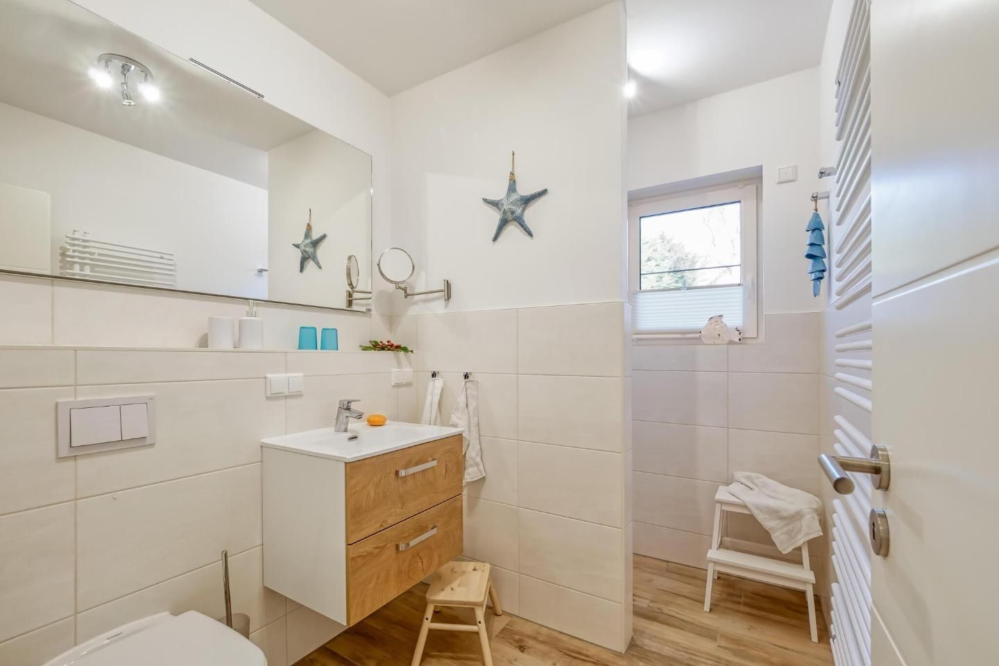 A modern bathroom with a white vanity, a small wooden stool, a mirror with star decorations, and a window with blinds.