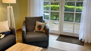 Een woonkamer met een bank, fauteuil en een vloerlamp. Er is een groot raam met uitzicht op groen.