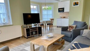 Een woonkamer met een grijze bank, een houten salontafel en een televisie. Er is een eethoek met een tafel en stoelen, en een keuken op de achtergrond.