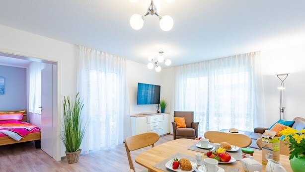 Quarto de hotel moderno com uma cama, área de refeições com mesa posta para o café da manhã, televisão e uma grande janela com cortinas.