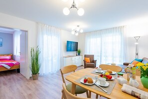 Moderni hotellihuone, jossa on sänky, ruokailualue aamiaispöydällä, televisio ja suuri ikkuna, jossa on verhot.