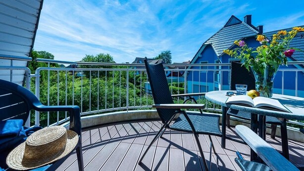 Een houten terras met een ronde tafel en stoelen, een rieten mand en een vaas met bloemen.