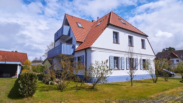 Une maison à deux étages, avec un toit recouvert de tuiles rouges, un balcon à l’étage supérieur et un petit jardin à l’avant.