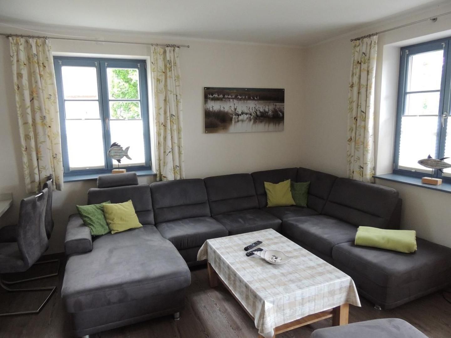 Ein Wohnzimmer mit einem grauen Ecksofa, einem kleinen Tisch mit kariertem Tischdecken und zwei mit Vorhängen versehenen Fenstern.