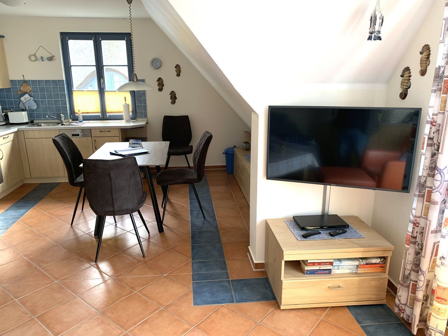 A compact kitchen with a dining area, a TV on a stand, and a small bookshelf.