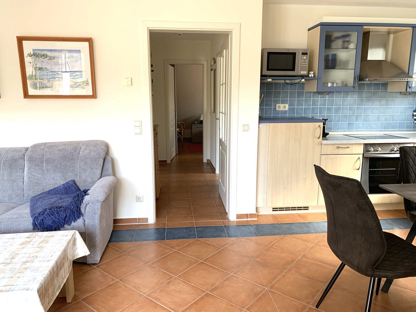 A living room with a grey sofa, a blue throw pillow, and a dining table with a chair. A kitchen with a microwave, oven, and blue-tiled backsplash is visible in the background.