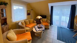Una sala de estar acogedora con muebles de mimbre, una mesita con un objeto decorativo y vistas al exterior a través de amplios ventanales.