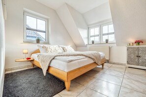 Quarto com teto inclinado, cama com estrutura de madeira, mesa de cabeceira com abajur e cômoda com painéis decorativos.