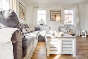 Ein modernes Wohnzimmer mit einer grauen Eckcouch, einem weißen Couchtisch und einer Stehlampe. Zwei Fenster mit Vorhängen, ein Heizkörper und ein gerahmtes Bild sind ebenfalls vorhanden.