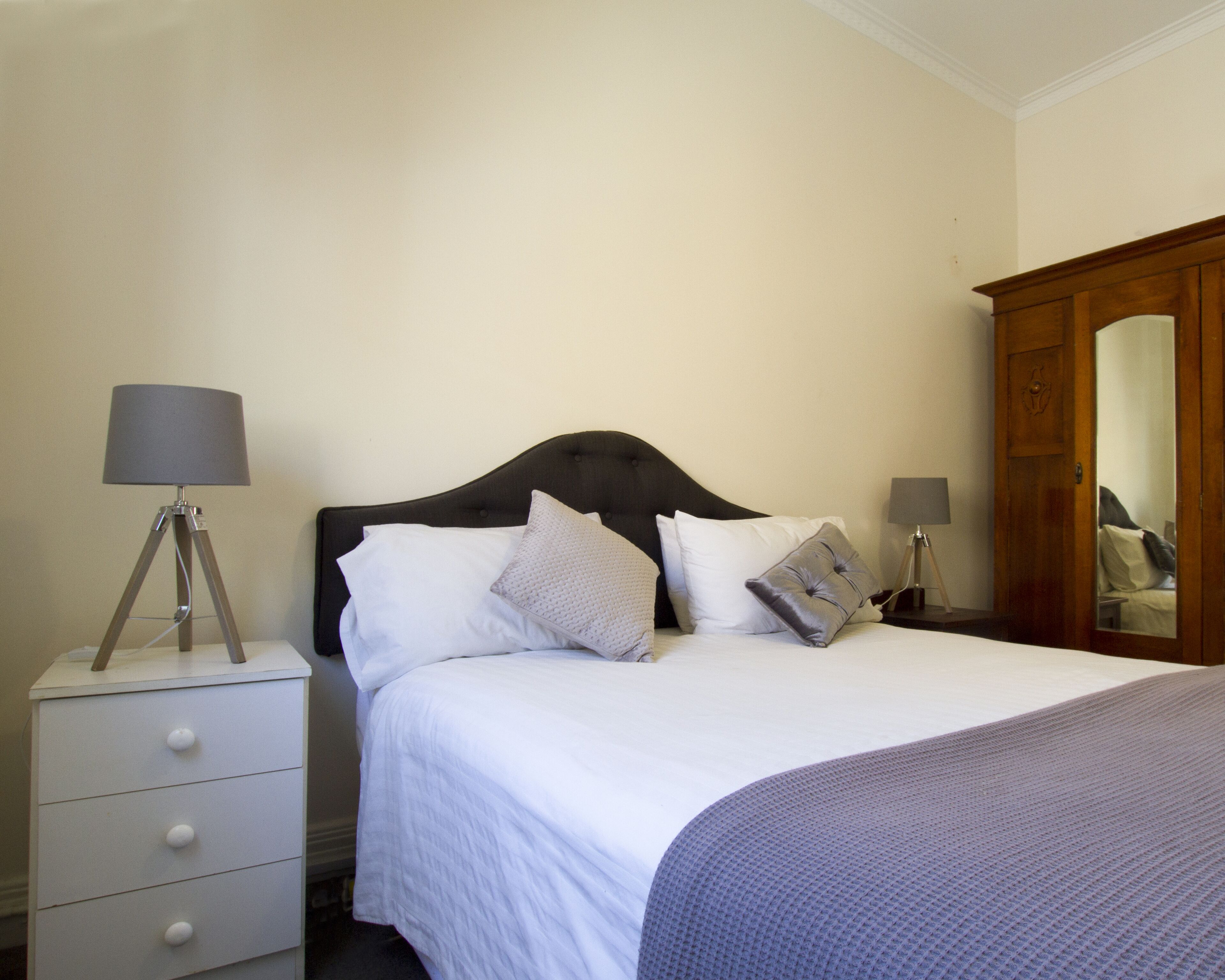 A neatly made bed with a dark headboard, white bedding, and several pillows. There is a bedside table with a lamp and a small drawer. A wooden wardrobe is visible to the right.
