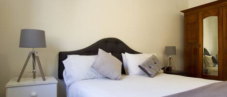 A neatly made bed with a dark headboard, white bedding, and several pillows. There is a bedside table with a lamp and a small drawer. A wooden wardrobe is visible to the right.