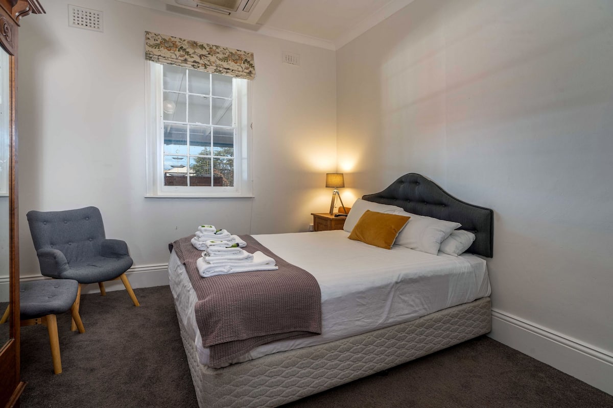 A bedroom with a bed, a chair, a nightstand with a lamp, and a window with a view.