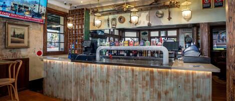 Una zona de bar con una barra de madera rústica, luces colgantes y un televisor que muestra un noticiero.