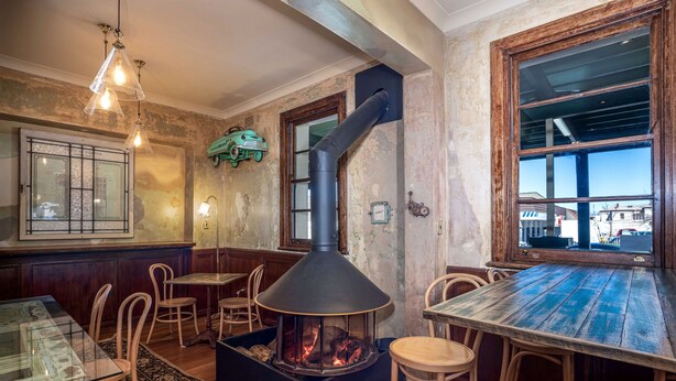 Un interior rústico con un horno de leña central, mesas y sillas de madera, y un amplio ventanal con vistas.