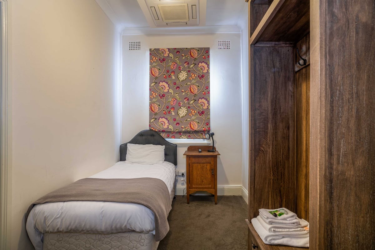 A small bedroom with a bed, a nightstand, a window with floral curtains, and a wooden wardrobe.