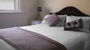 Una cama bien tendida con colcha a rayas y una manta marrón, una lámpara de noche y una ventana con cortina estampada.