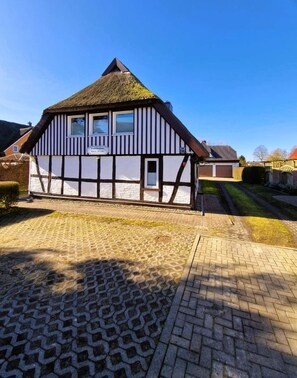A traditional half-timbered house with a thatched roof, a paved driveway, and a clear blue sky.