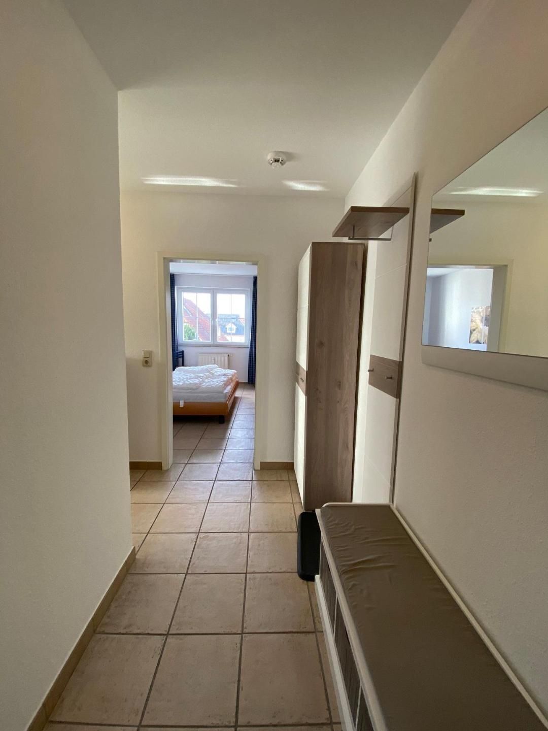 A narrow hallway with a bed in the background, a wooden wardrobe, and a mirror on the wall.