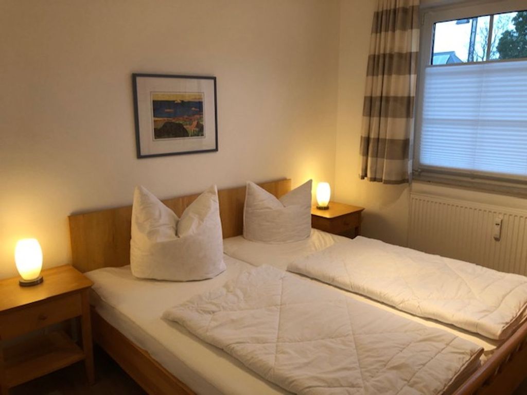 Ein Doppelbett mit weißer Bettwäsche, zwei Nachttische mit Lampen, ein gerahmtes Bild an der Wand und ein Fenster mit Vorhängen.