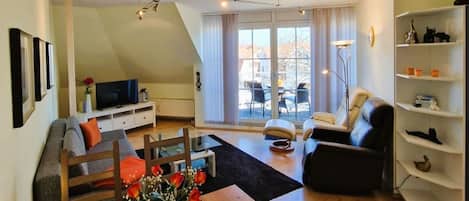 Ett modernt vardagsrum med ett matbord i trä, en soffa, en svart fåtölj och en bokhylla.