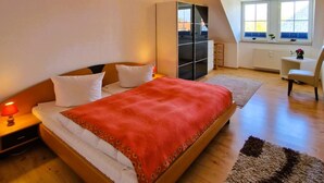 Ein Schlafzimmer mit Bett, Nachttischen, Kommode, Stuhl, Fenster und Heizkörper.