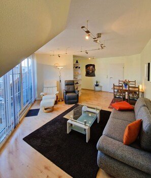 A spacious living area with a sofa, armchair, and coffee table, a dining table with chairs, a bookshelf, and a large window. - Strandstrasse 51 Whg. 16 - Modern and cozy vacation apartment (Zingst)