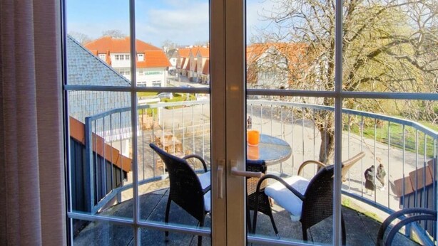 Ein Zimmer mit großem Fenster, Blick auf ein Gebäude mit rotem Dach und einem Balkon mit Stühlen und Tisch.