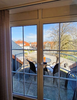 Ein Zimmer mit großem Fenster, Blick auf ein Gebäude mit rotem Dach und einem Balkon mit Stühlen und Tisch.