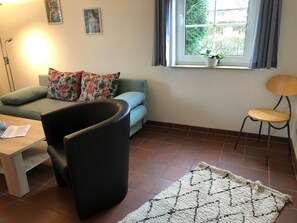 Uma sala de estar com sofá, poltrona, mesa pequena, janela com cortinas e tapete.