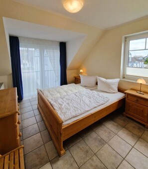 Une chambre à coucher avec un lit en bois, une table de chevet, une commode, une fenêtre avec des rideaux et une vue sur l’extérieur.