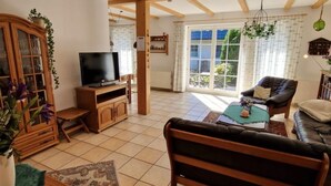 Een woonkamer met een houten bank, een televisie op een houten dressoir, een kast met glazen deuren en een groot raam met gordijnen.