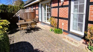 Een tweelaags huis met een bakstenen gevel, grote ramen en een verhard terras met tuinmeubilair.