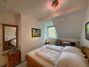 Ein Schlafzimmer mit Bett, Kommode, Spiegel, Fenster und Deckenleuchte.