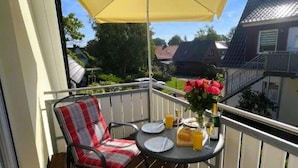 Un balcon avec un parasol jaune, deux chaises dotées de coussins à carreaux, une petite table sur laquelle sont disposés des éléments pour le petit-déjeuner, et un vase rempli de fleurs.