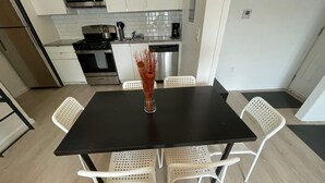 Une cuisine moderne avec des meubles blancs, un plan de travail noir et un réfrigérateur en acier inoxydable. On y trouve une table noire sur laquelle est posé un vase rempli de branches séchées, ainsi que quatre chaises blanches.
